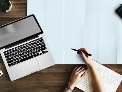 A person writing in a notebook with her laptop on the side